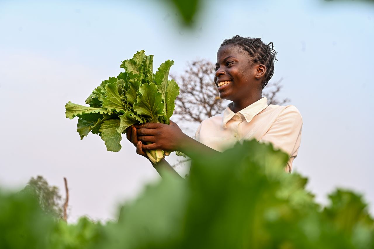 A young farmer smiles while holding a freshly cut bunch of leafy greens, framed by the crop around her.